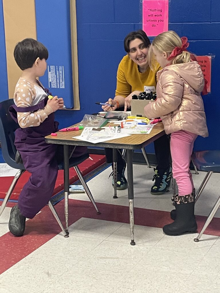 A Four Corners Project team member in a yellow sweater engages two young children at a table covered with markers and drawing materials during a community design workshop.