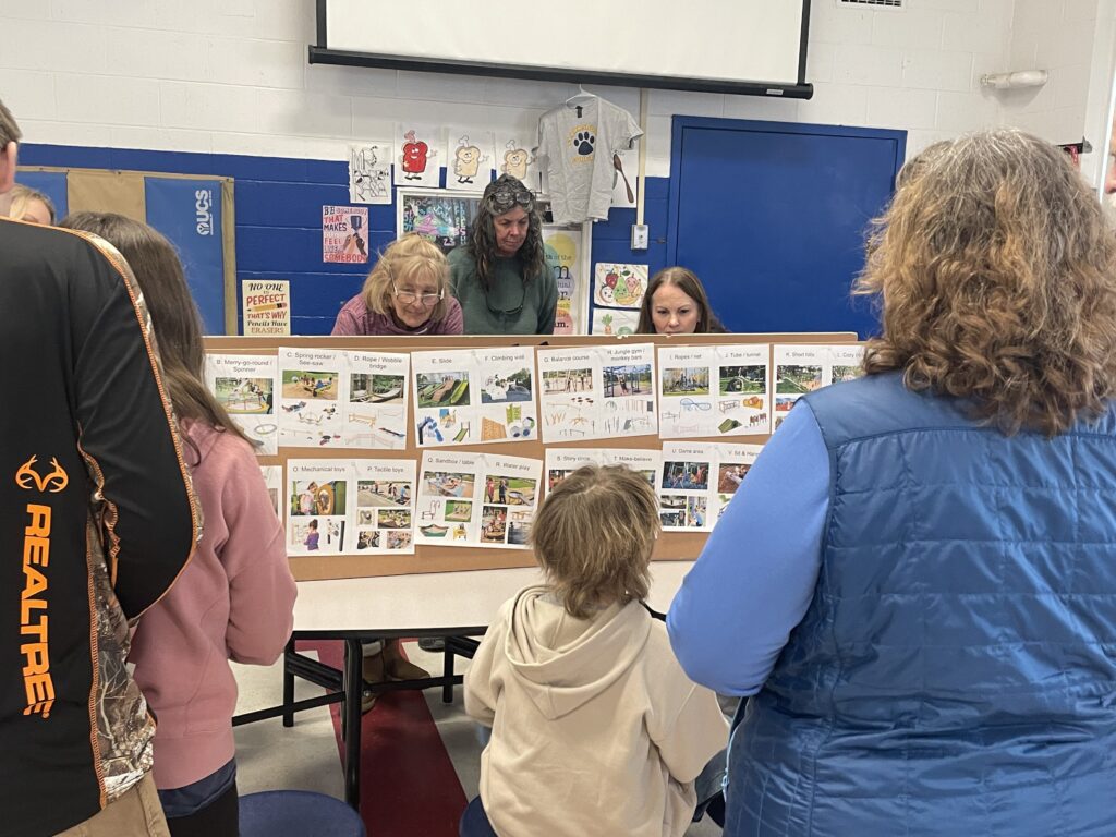 Adults and children gathered around a display board covered with labeled photographs of playground equipment, reviewing options during a community design engagement session.